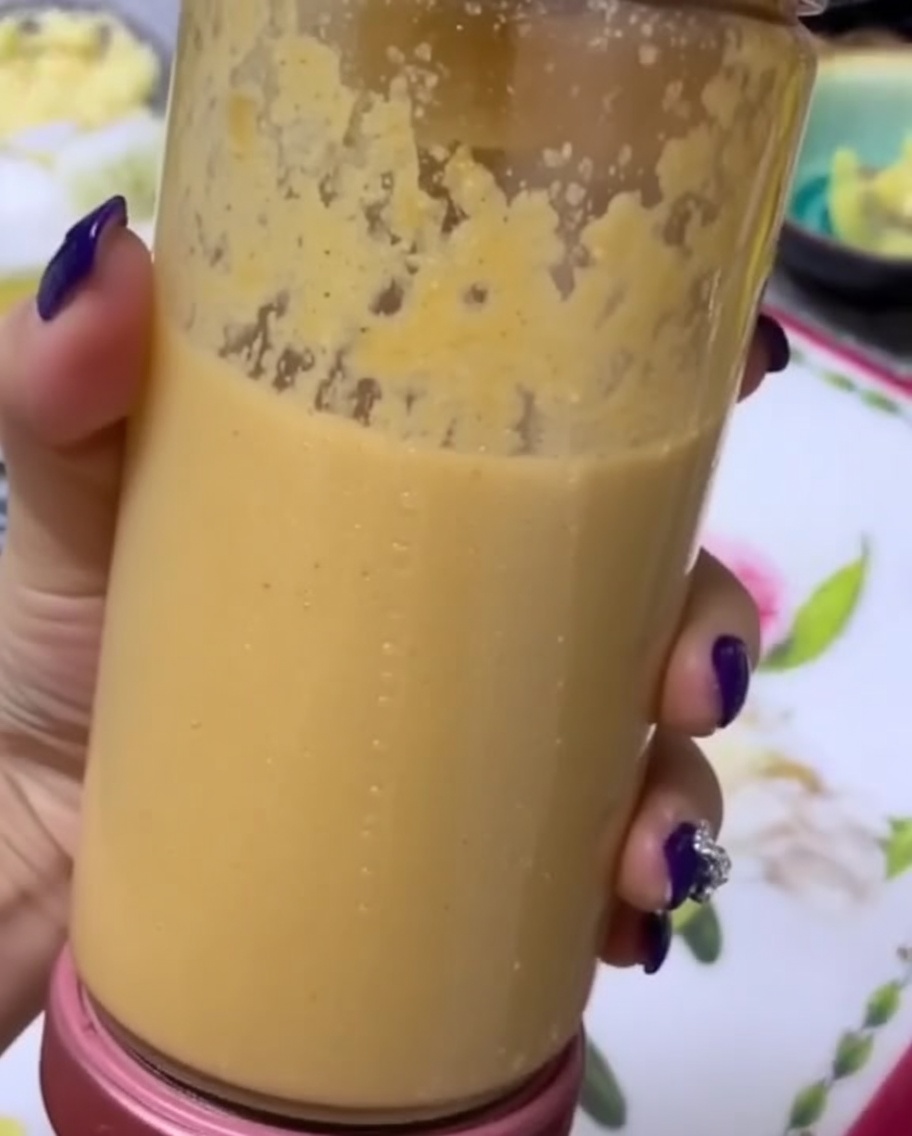 A hand holding a clear blender canister filled with a smooth, thick fruit and vegetable puree.