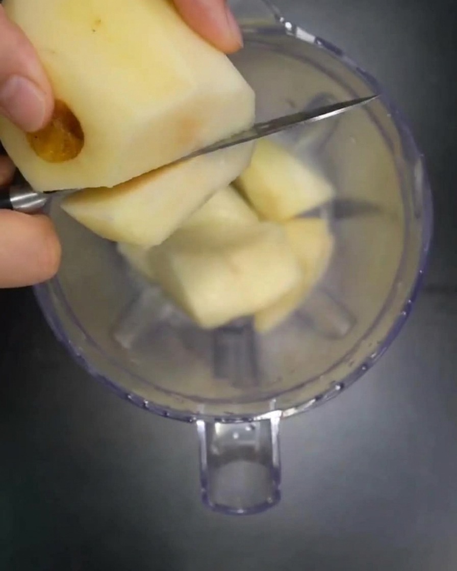 Chunks of fresh pear, apple, and onion being dropped into a clear blender pitcher.