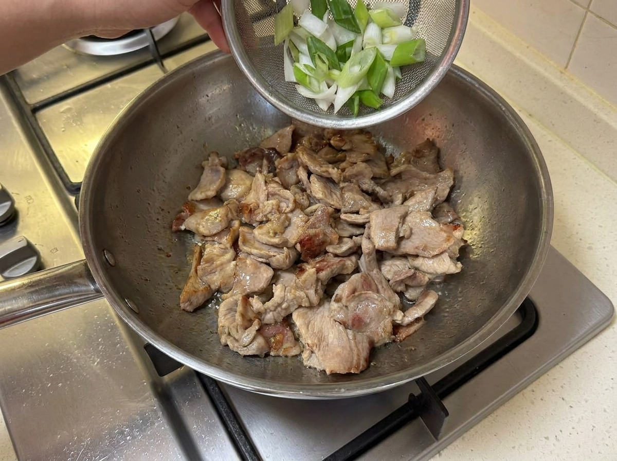 Sliced green onion segments being dropped into the pan with the browned pork.