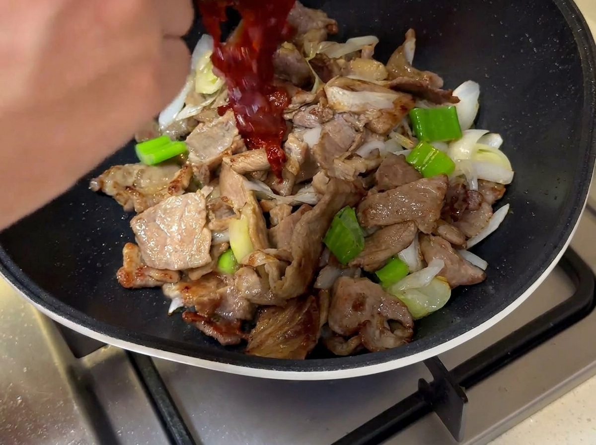 The dark red sauce being poured from a small bowl into the pan over the meat and onions.