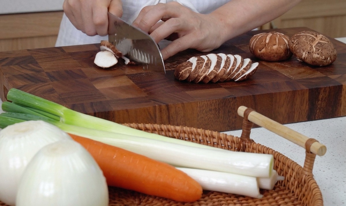 Hands using a large kitchen knife to slice fresh shiitake mushrooms on a wooden cutting board.