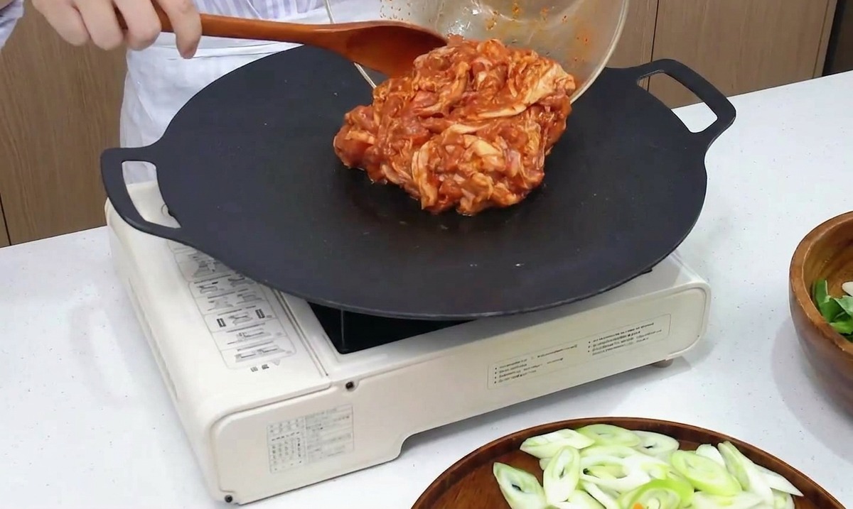 Marinated spicy pork belly being poured from a glass mixing bowl onto a hot black griddle pan.