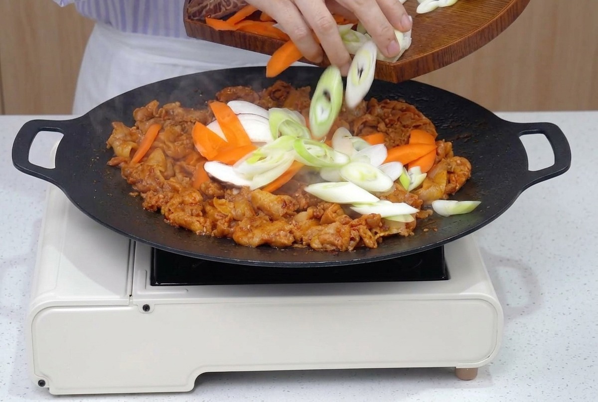 A pan filled with browned pork belly being joined by fresh slices of carrots, onions, mushrooms, and green onions.