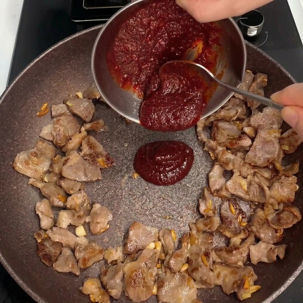 Thick red sauce being poured into the center of the pan with stir-frying pork.