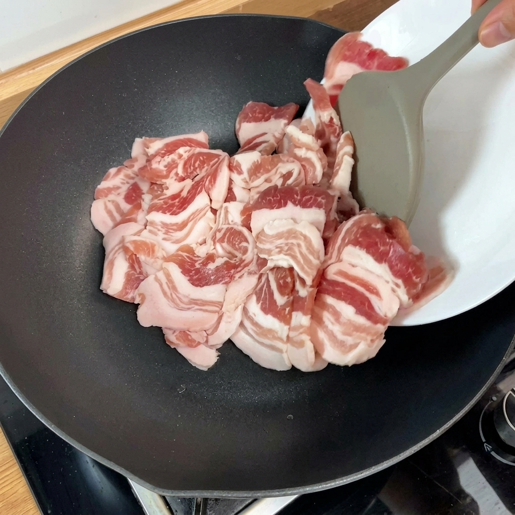 Slices of raw pork belly being added to a hot pan with a kitchen tool, showing the start of the searing process.