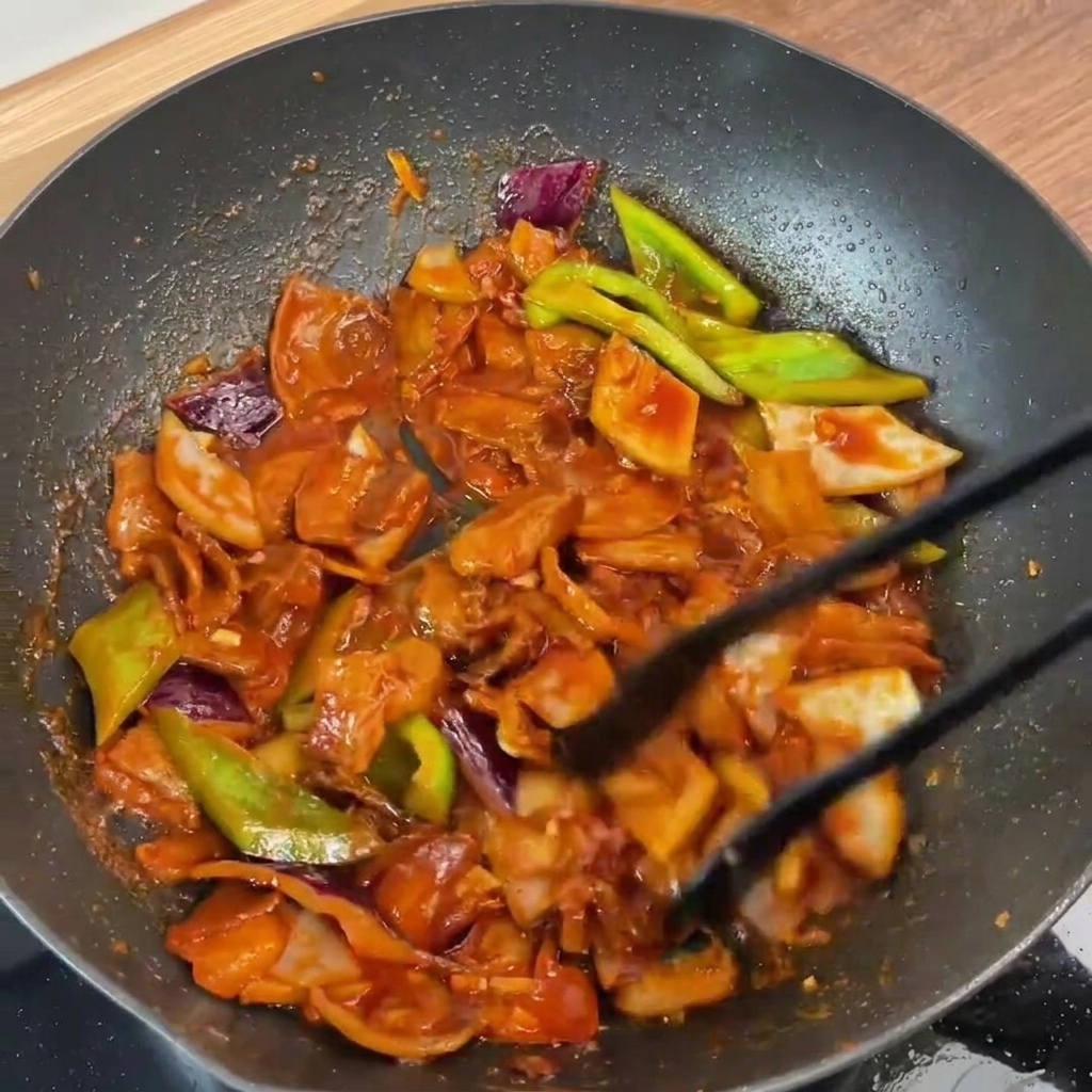 The finished stir-fried pork belly and vegetables being tossed together in the pan with tongs, perfectly coated in the glossy red sauce.