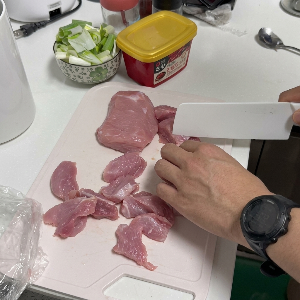 A person slicing raw pork on a white plastic cutting board with a white kitchen knife.