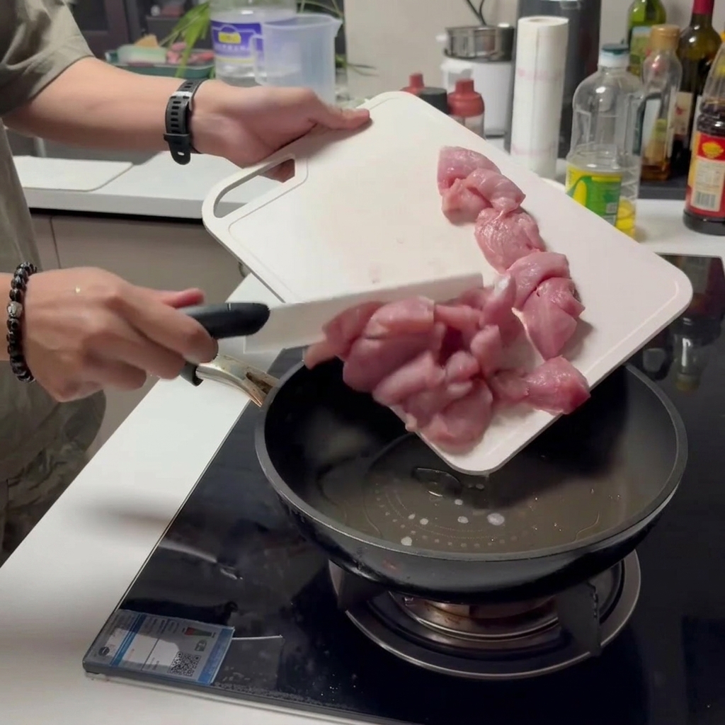 Sliding pieces of raw sliced pork off a white cutting board directly into an oiled frying pan.