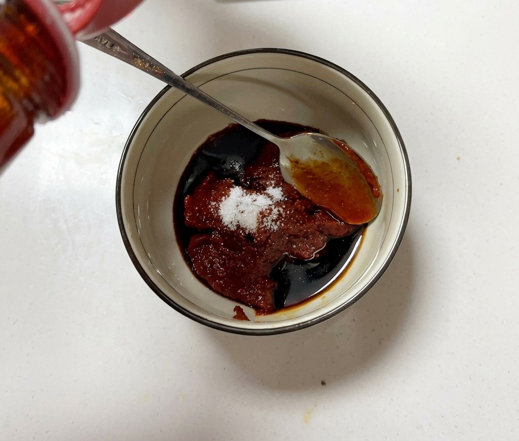 A small bowl filled with Korean chili paste, sugar, and soy sauce being mixed with a metal spoon.