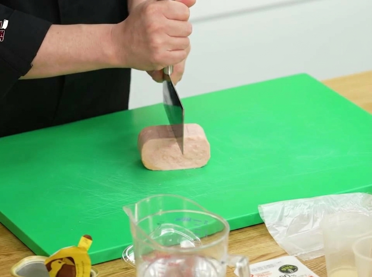 A block of Spam being sliced in half with a large kitchen knife on a green cutting board.