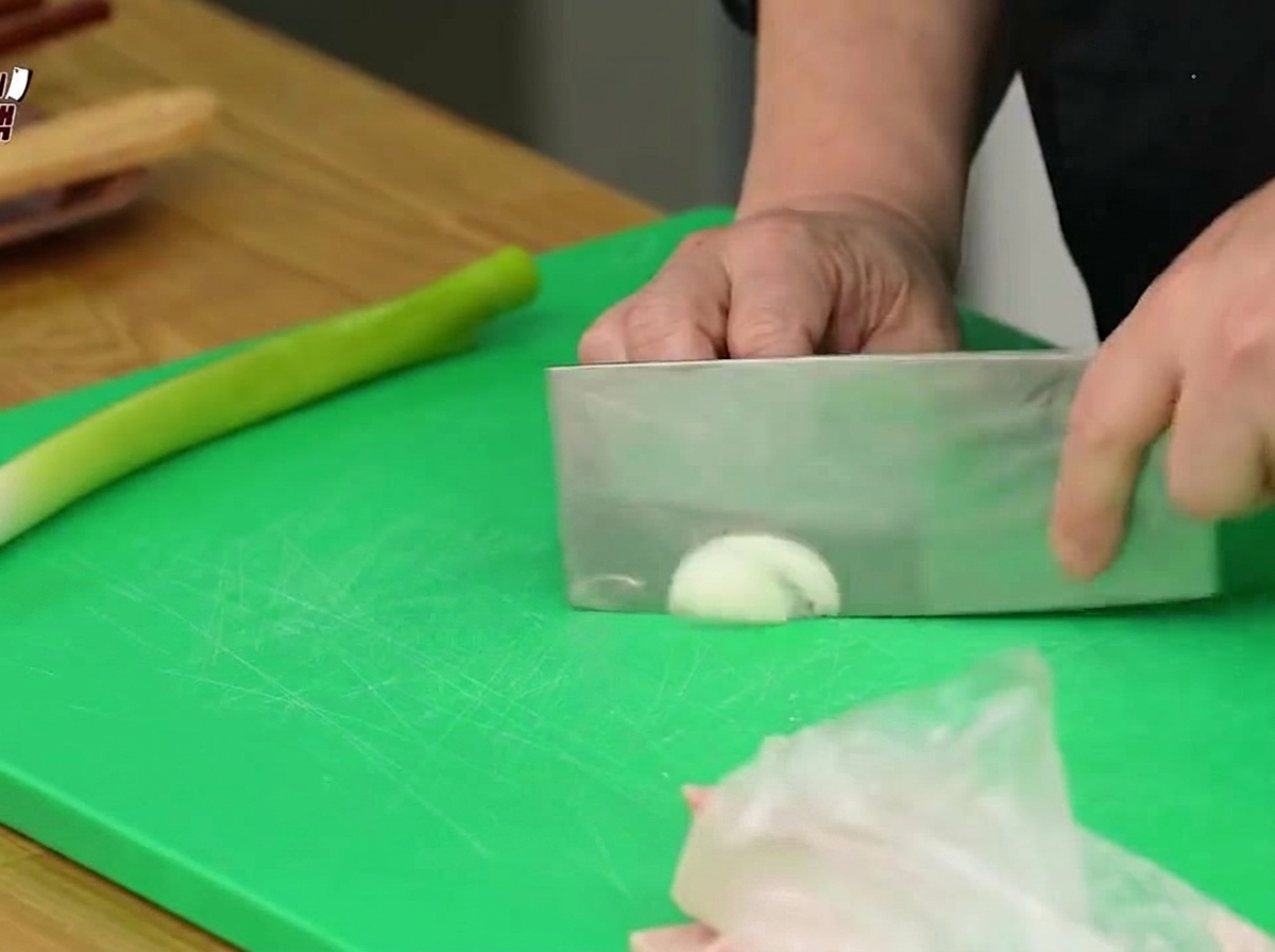 An onion being sliced into thin strips on a green cutting board using a cleaver.