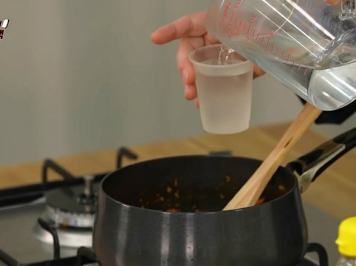 Water being poured from a measuring cup into a black cooking pot containing sautéed ingredients.