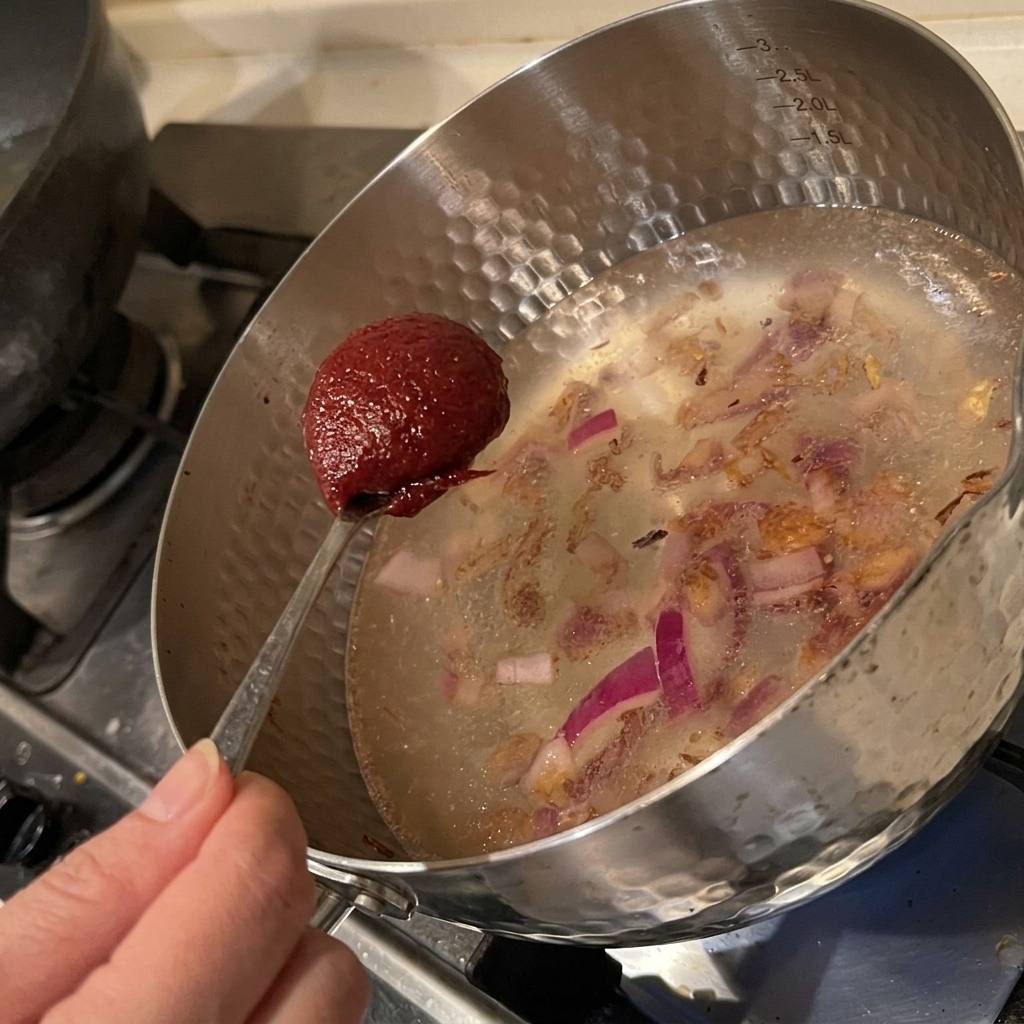 A spoonful of red Korean chili paste being added into a pot of water and sautéed onions.