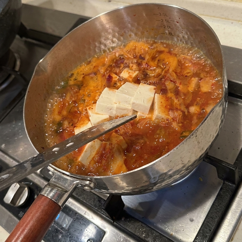 A variety of fresh ingredients including tofu blocks, enoki mushrooms, and kimchi being simmered in a spicy, red-tinted broth.