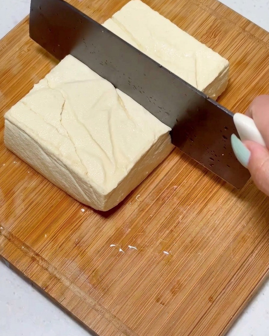 Slicing a square block of firm tofu into uniform pieces using a large knife on a wooden surface.