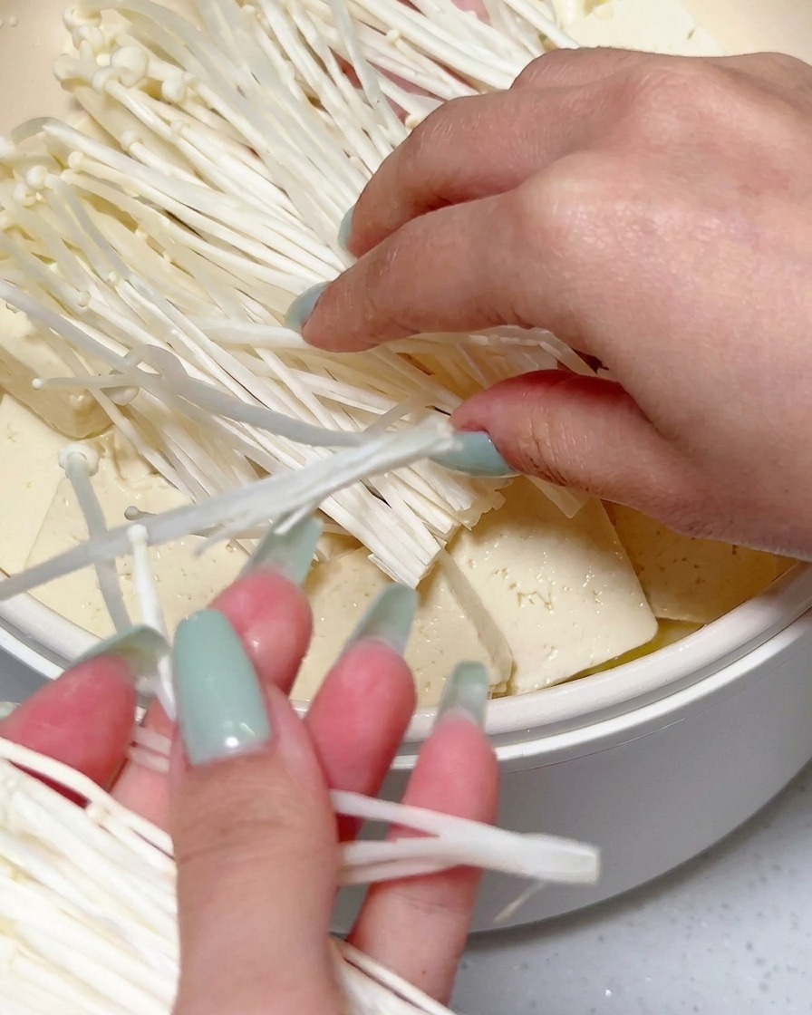 Hands placing a large cluster of fresh white enoki mushrooms on top of sliced tofu and other ingredients in a cooking pan.