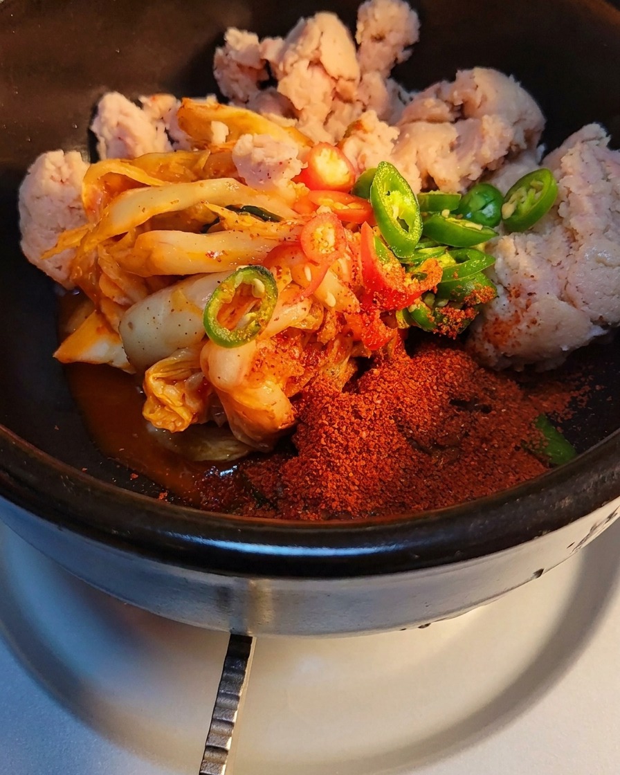 Close-up of sugar and beef seasoning powder being sprinkled over kimchi and luncheon meat in a stone pot.