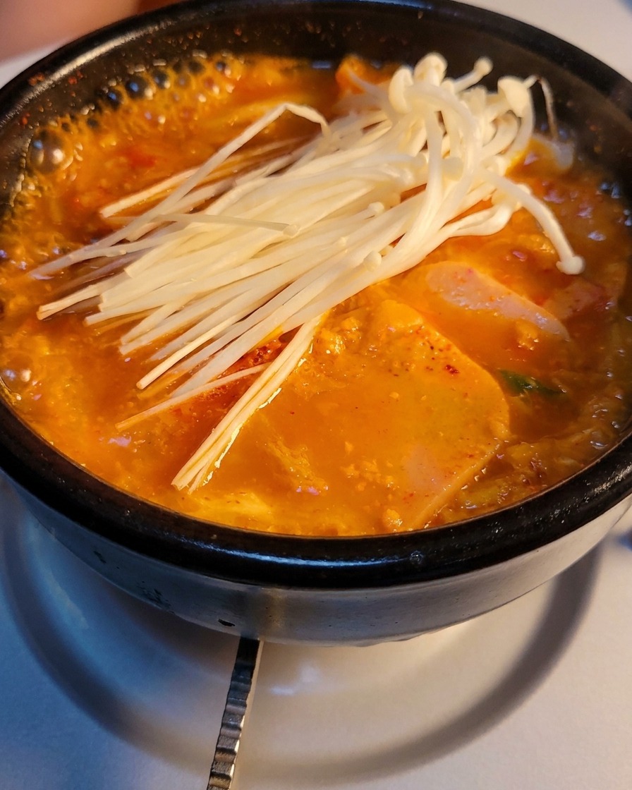 A stone pot of bubbling kimchi and luncheon meat soup topped with a bunch of fresh enoki mushrooms.