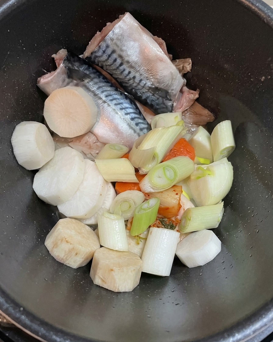A top-down view of a dark pot containing raw mackerel fillets, chunky white radish pieces, and fresh sliced green onions.