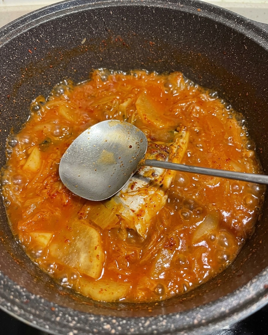 A vibrant, bubbling red stew of mackerel and vegetables in a pot, with a metal spoon lifting a piece of fish.