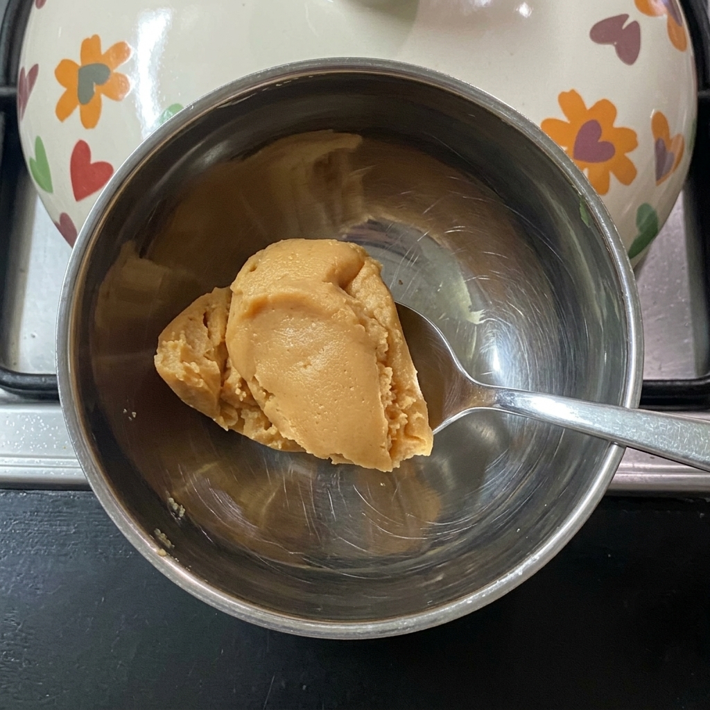 A spoon stirring miso paste into warm water in a small metal bowl.