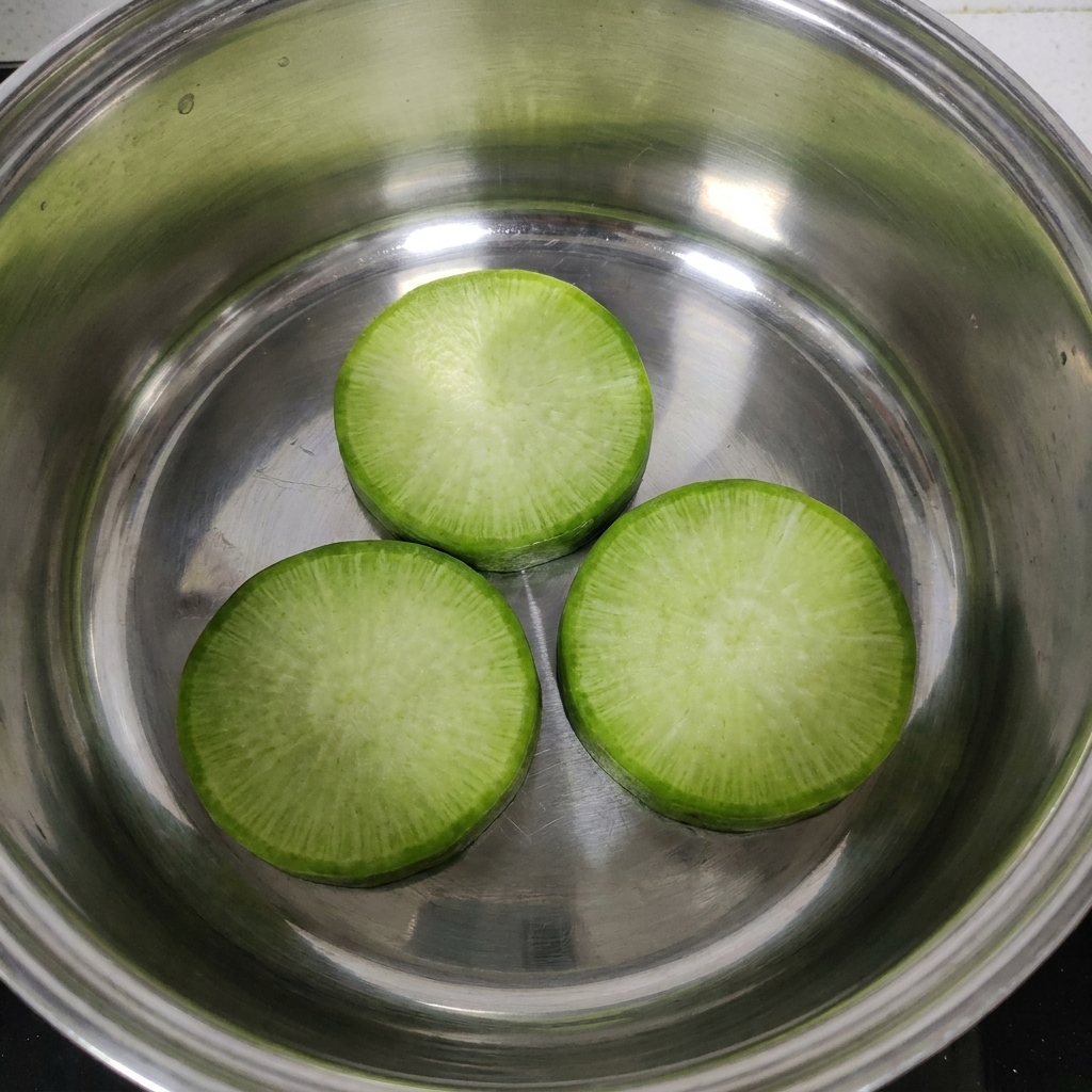 Three thick radish slices neatly placed at the bottom of a stainless steel cooking pot.