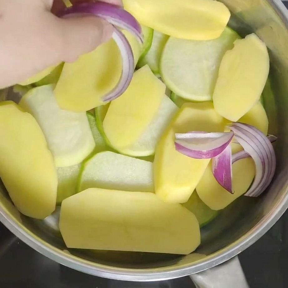Hand adding fresh red onion chunks into the pot on top of the potatoes.