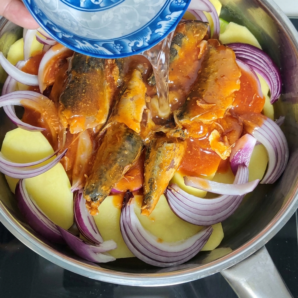 Water being poured from a small blue and white ceramic bowl into the pot containing mackerel and vegetables.