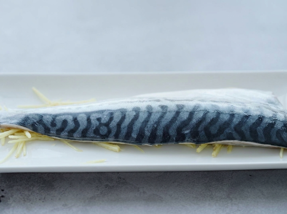 A raw mackerel fillet laid on top of a bed of thin ginger strips, with cooking sake being poured over it.