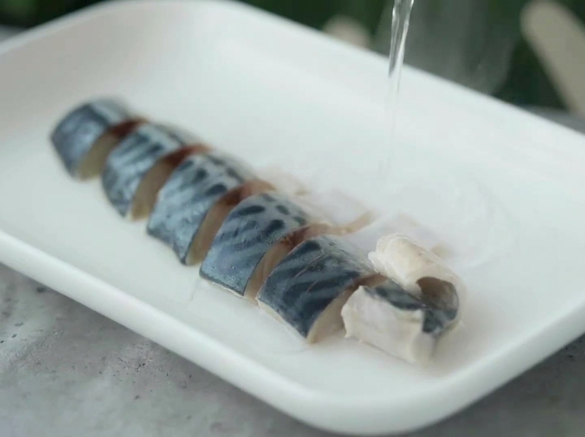 Hot water being poured from a kettle over arranged mackerel pieces on a white plate.