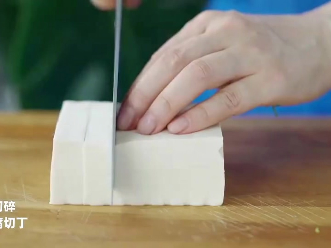 A chefs knife cutting a block of white tofu into precise, even-sized cubes on a wooden surface.