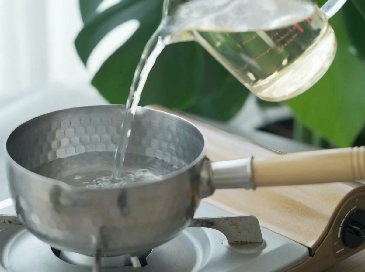 Clear dashi broth being poured from a measuring cup into a small stainless steel pot.
