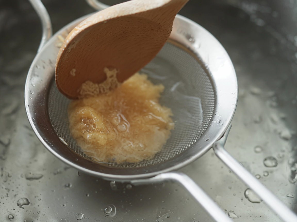 White miso paste being stirred through a metal strainer into boiling soup with a wooden spoon.