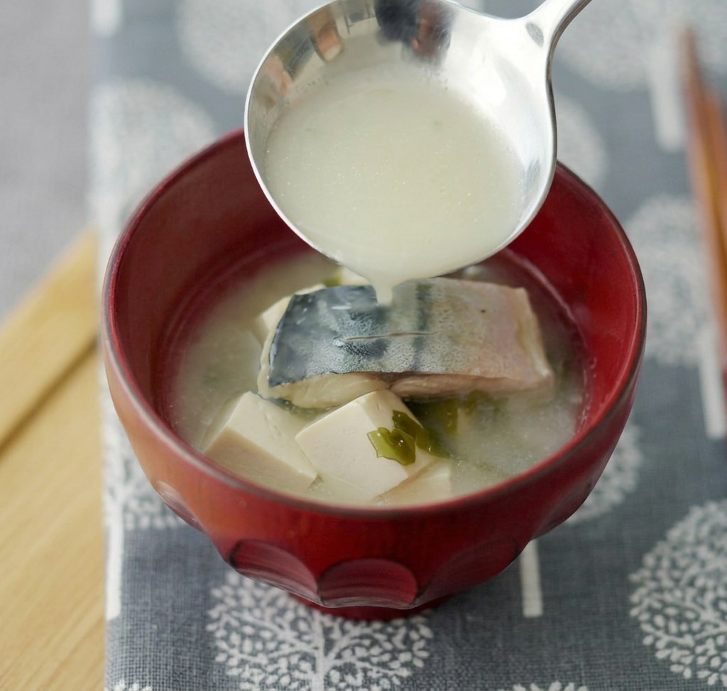 Miso soup with mackerel and tofu being ladled into a deep red ceramic bowl.