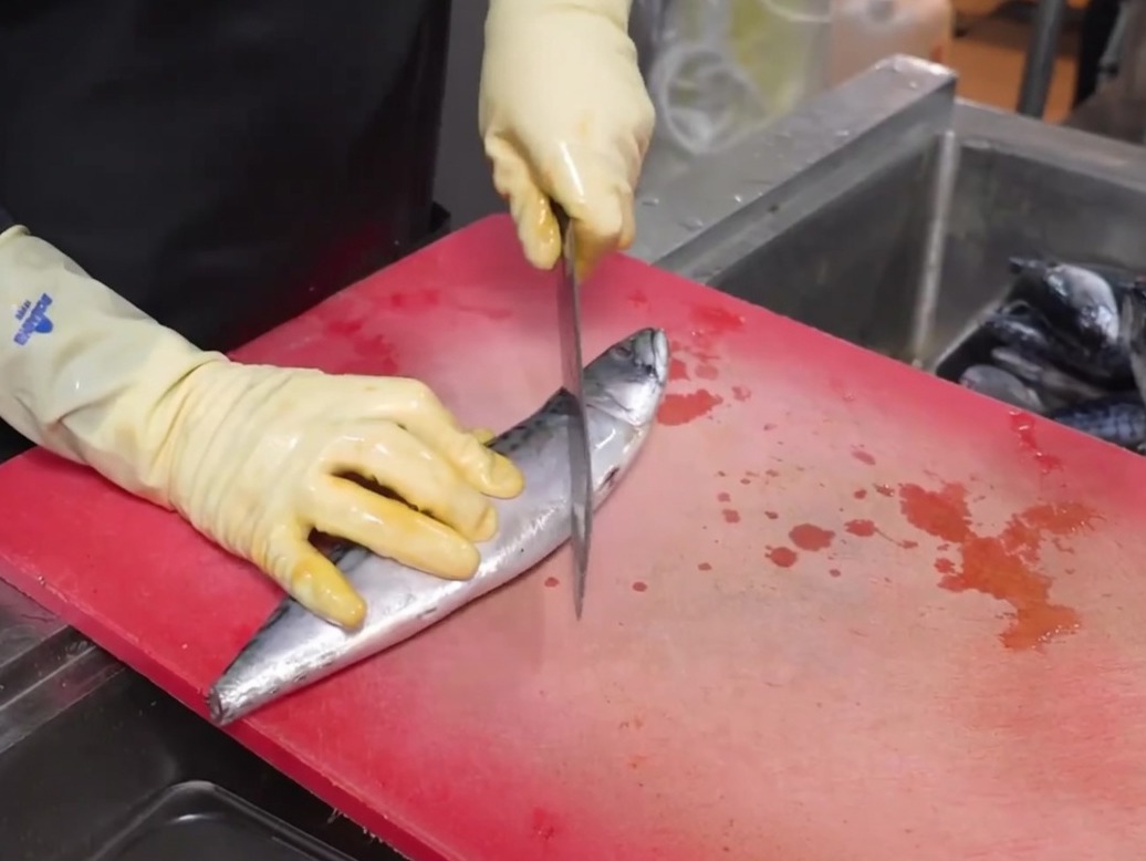 A knife making a clean cut through a whole mackerel on a red cutting board.
