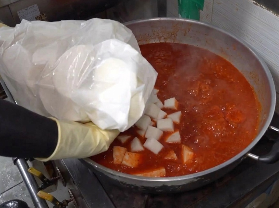 Radish chunks being added to a large pot filled with boiling spicy red liquid.