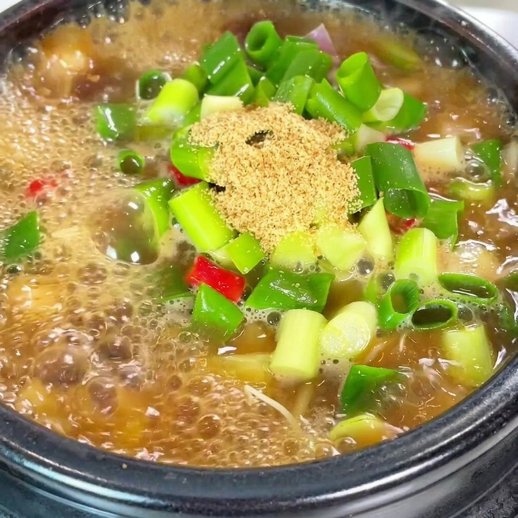 A close-up view of beef seasoning powder and bright red chili flakes being added into a boiling pot of soybean paste stew with scallions.