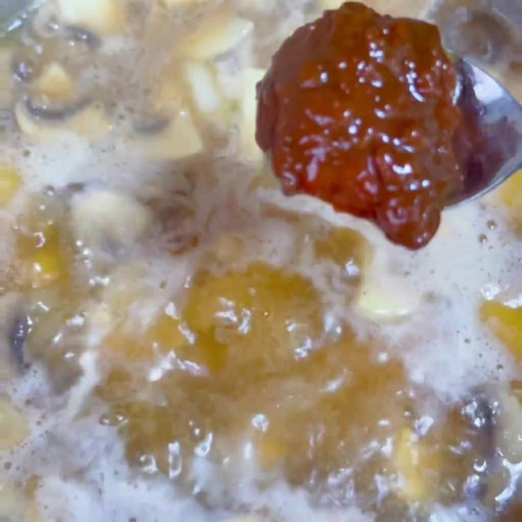 A spoonful of rich, dark soybean paste being stirred into a bubbling, savory broth in a wok.