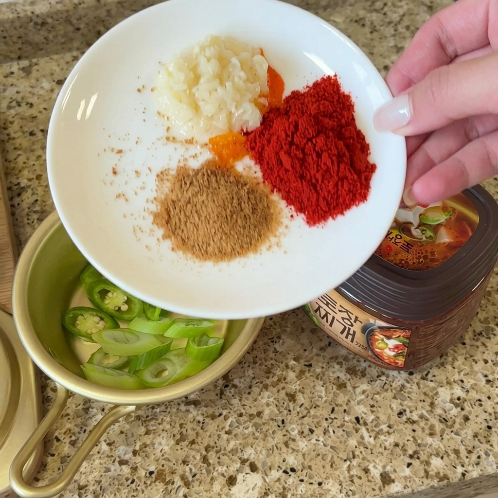 A white plate holding small portions of minced garlic, chili powder, beef bouillon, and perilla powder beside a container of doenjang.