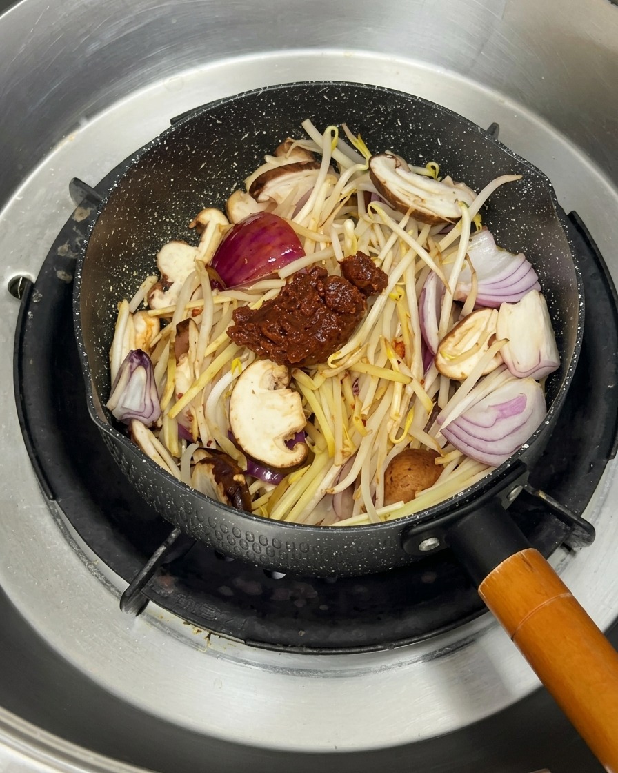 Boiling water being poured into a pan filled with stir-fried vegetables and Korean bean paste.