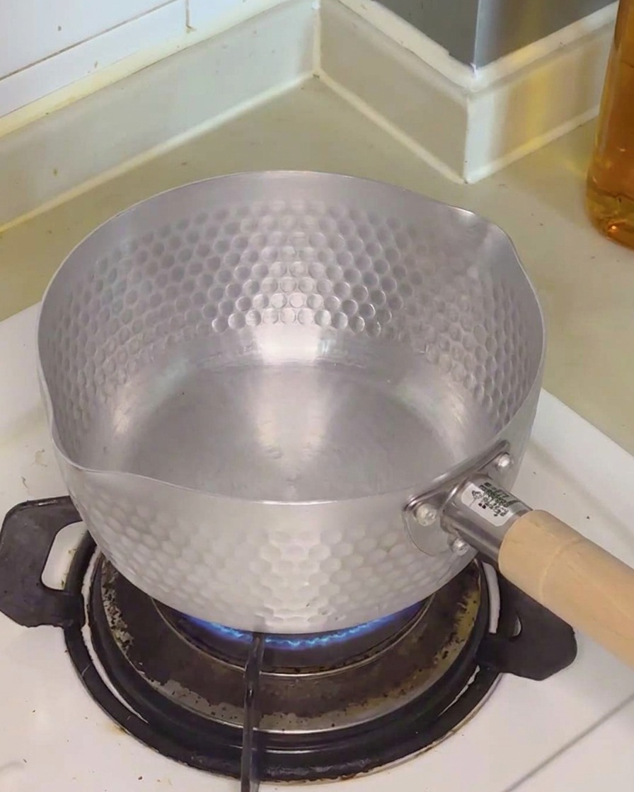 Clear water being poured into a small, hammered-finish metal pot sitting on a gas stove burner.