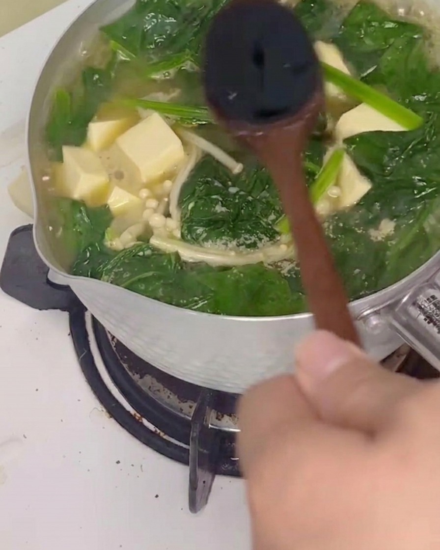 Soy sauce being stirred into the bubbling spinach and tofu soup with a wooden spoon.