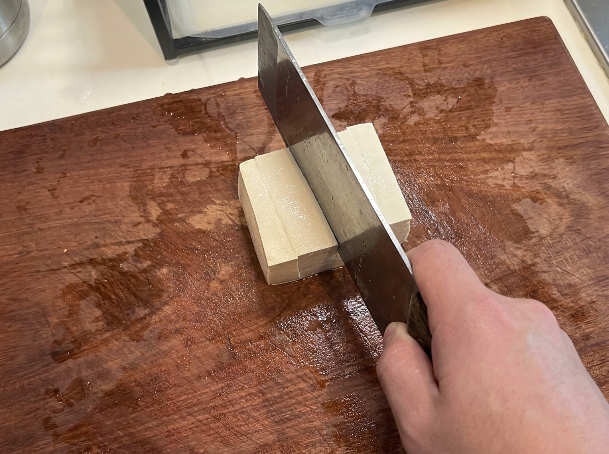 A block of firm tofu being cut into neat, uniform cubes on a cutting board.