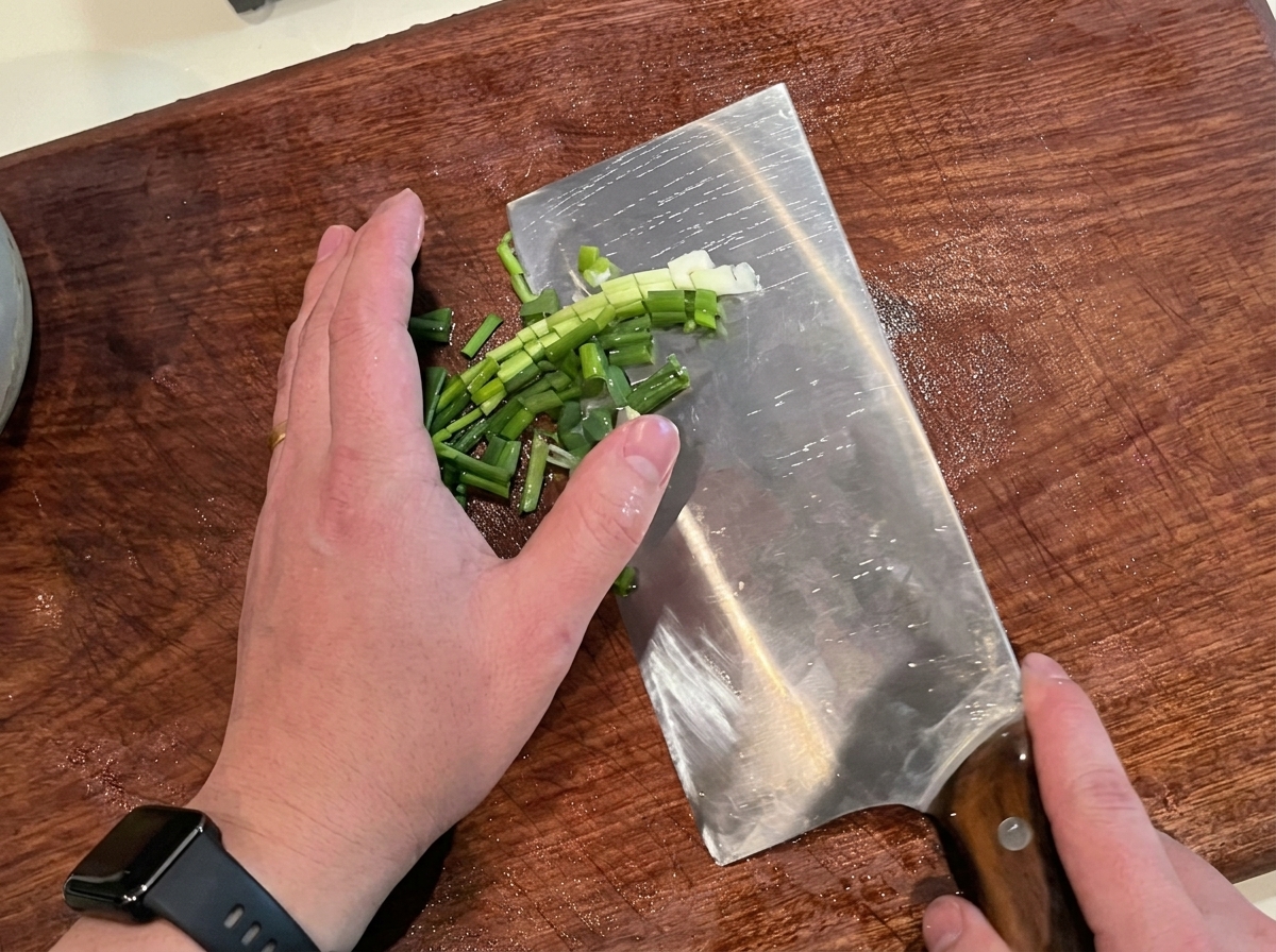 Fresh green scallions being finely chopped on a wooden cutting board.