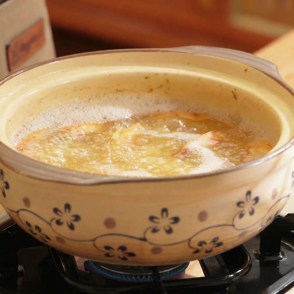 A clay pot filled with boiling, orange-tinted broth, with seafood and shiitake mushrooms being stirred in.