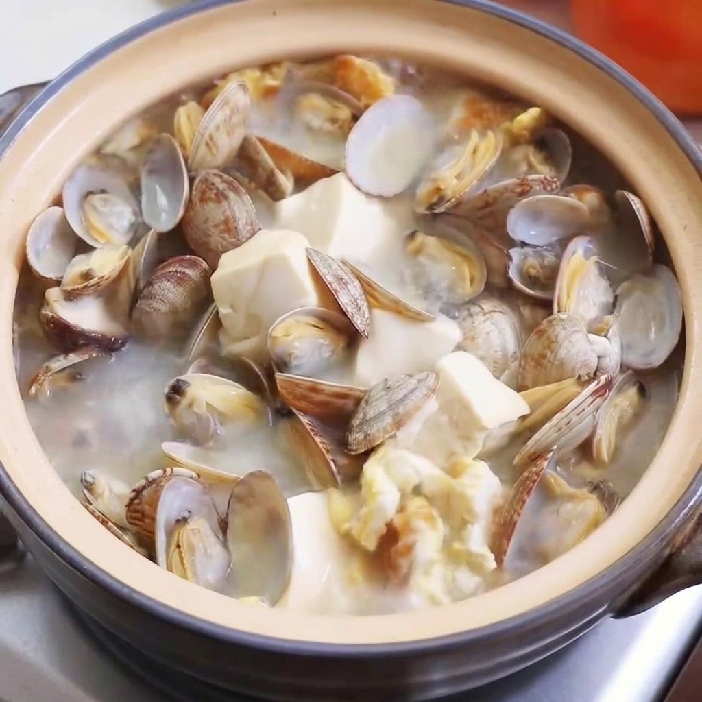 Close-up view of chicken powder, salt, and pepper being sprinkled into the bubbling clam and tofu soup.