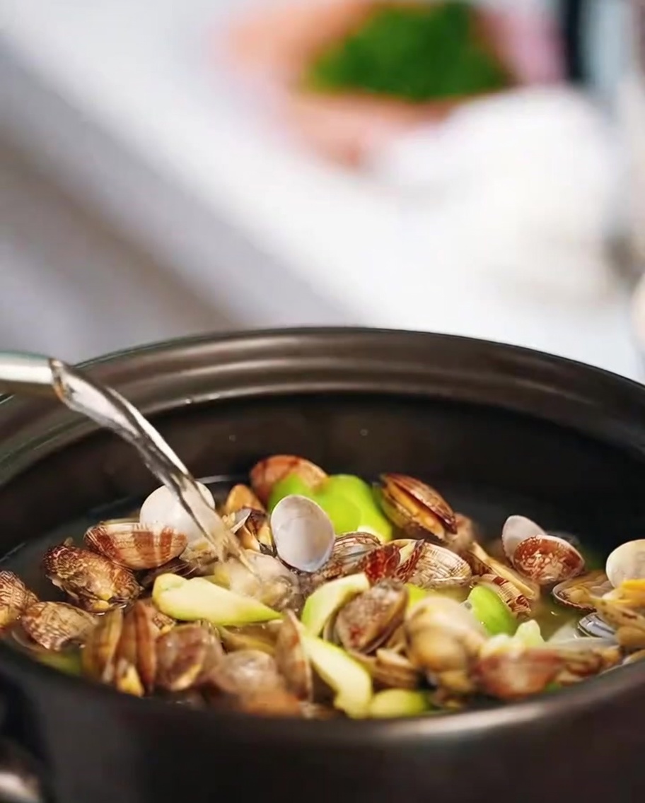 Hot water being poured into a clay pot filled with clams and luffa slices.