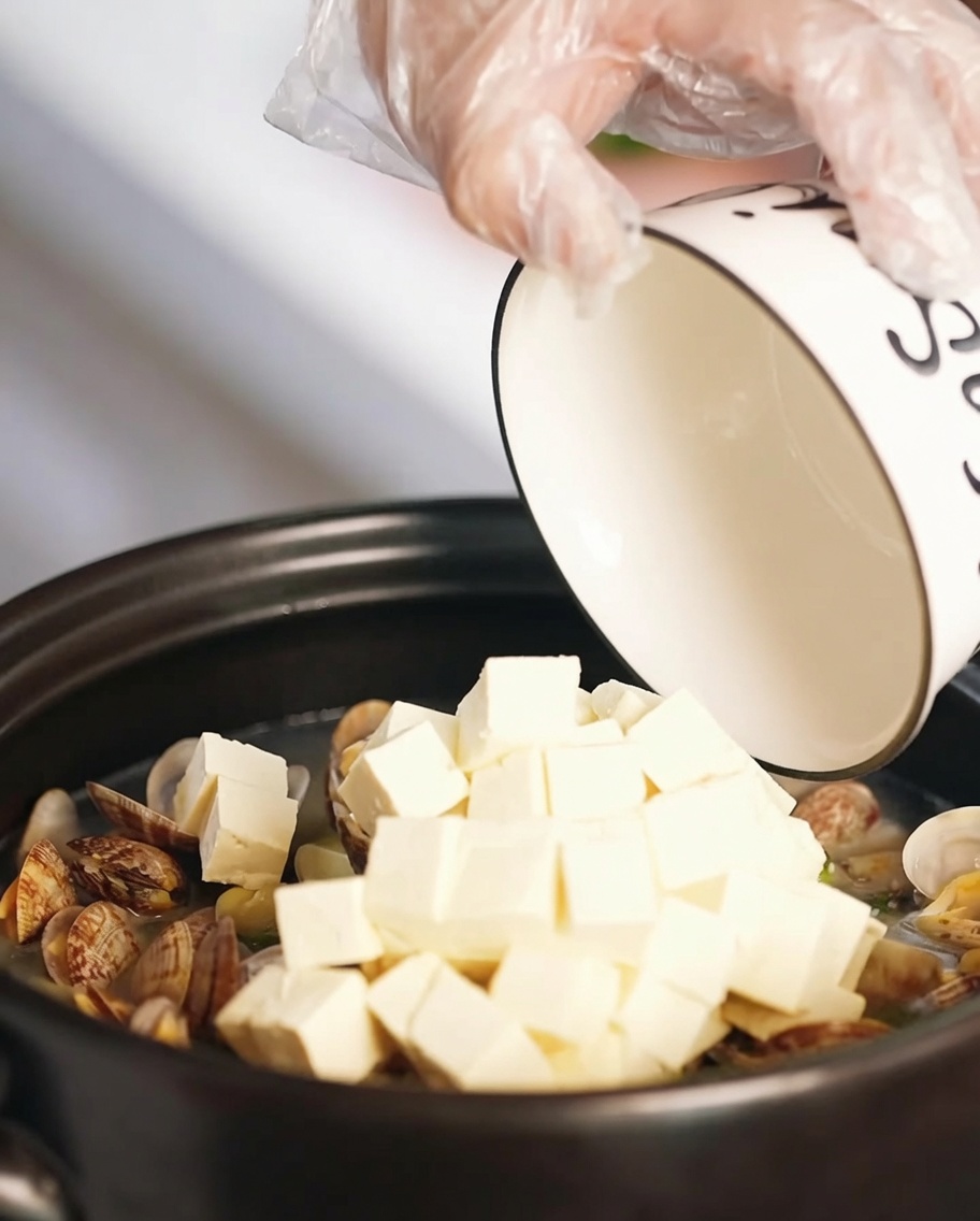 Soft tofu cubes and pieces of golden fried egg being added to the clam soup in a clay pot.