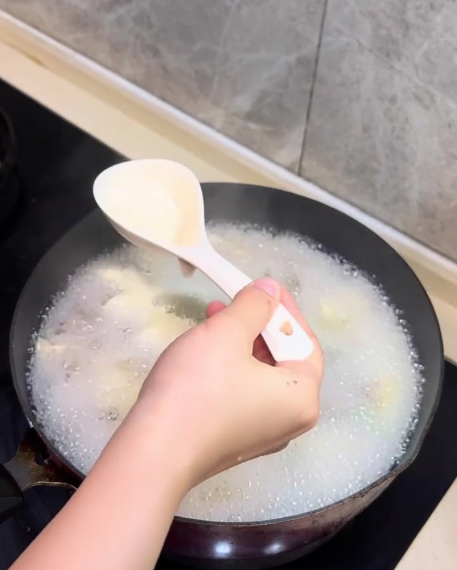 A spoon skimming white foam from the surface of the simmering clam and tofu soup.