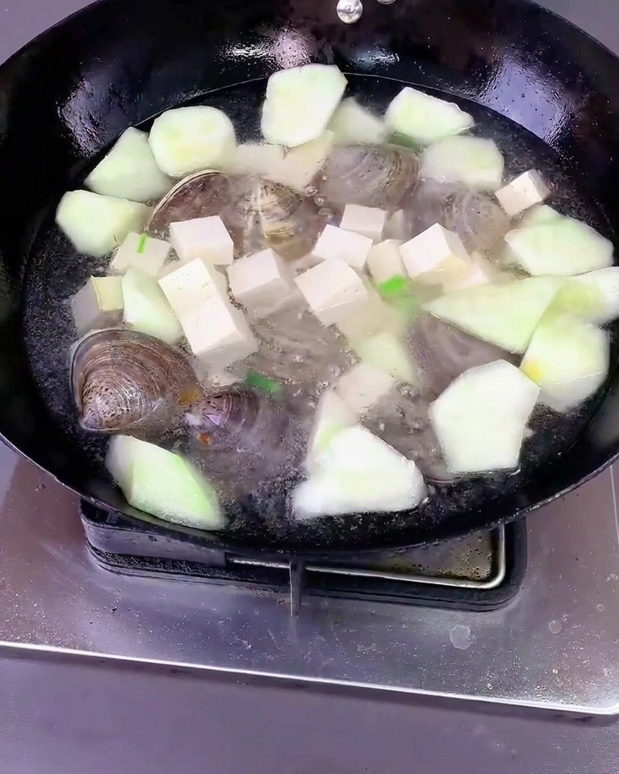 Water poured over luffa, tofu, and clams in a wok with aromatics and seasonings, covered with a lid.